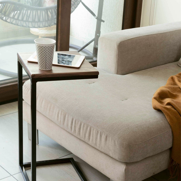 Mesa lateral en C con cubierta de madera tono nogal deslizada sobre un sofá beige, con una taza y una tablet