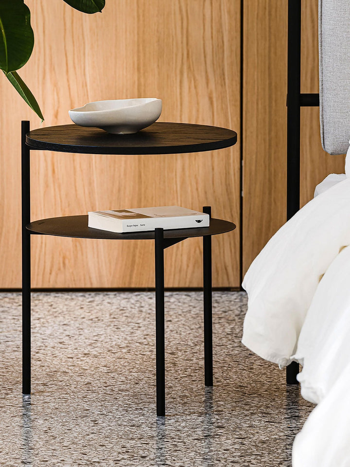 Velador de acero negro con cubierta de madera oscura junto a una cama, con un bowl de cerámica