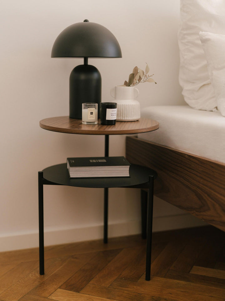 Velador con cubierta de madera en tono nogal junto a la cama, con lámpara negra y velas encima