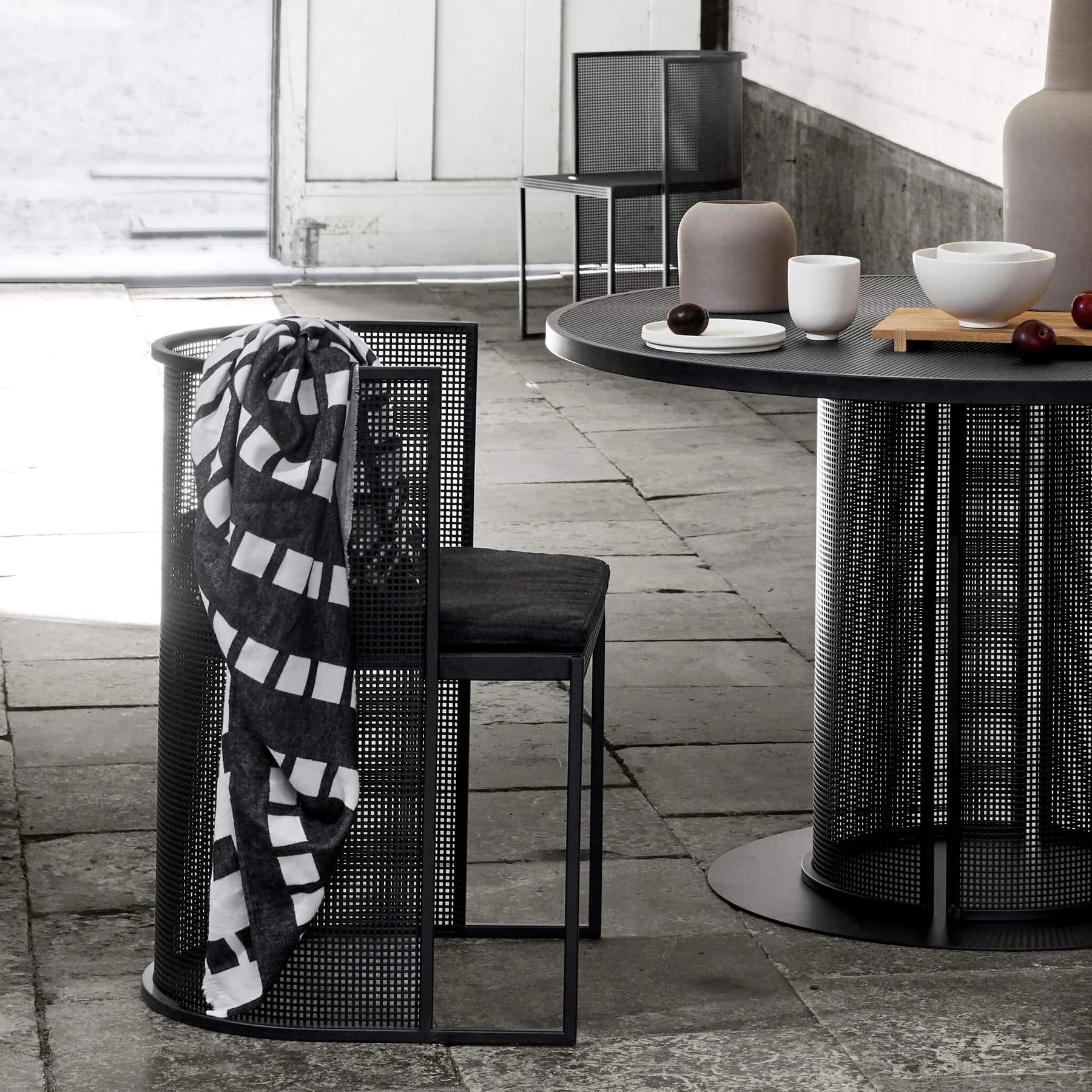 Silla de acero perforado negro con una manta sobre el respaldo, junto a una mesa redonda de acero
