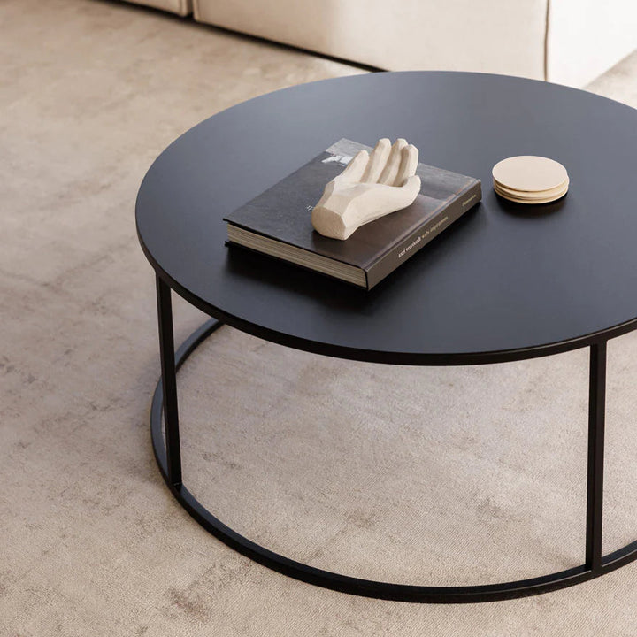 Mesa de centro circular de acero negro con un libro y una escultura de mano sobre la cubierta, junto a un sofá claro