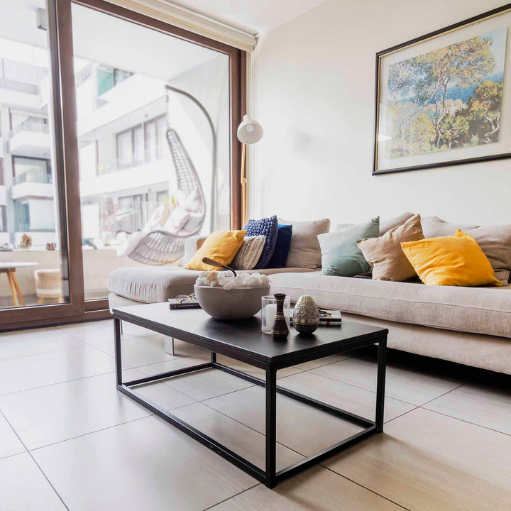 Mesa de centro rectangular con cubierta negra y estructura de acero, en un living con sofá beige y ventanal